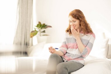 Mutlu genç kadın eliyle ağızdan hamilelik testine bakarken şaşkınlık ifade ediyor