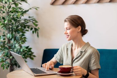 Kafede masa başında oturan, kahve içen ve dizüstü bilgisayar önünde web siteleri için net sörf casualwear meşgul genç kadın çalışan