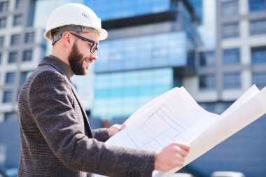 Takım elbiseli mutlu genç mühendis ve hardhat açık havada çalışırken plan üzerinde ne wbuilding kroki bakarak