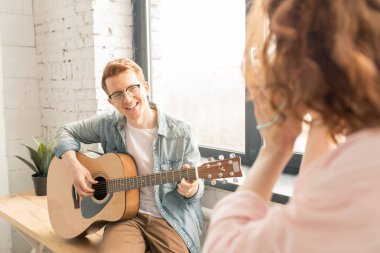 Mutlu genç şarkı adam ev parti kayıt kız tarafından düzenlenen akıllı telefon kamera bakarken gitar çalarken