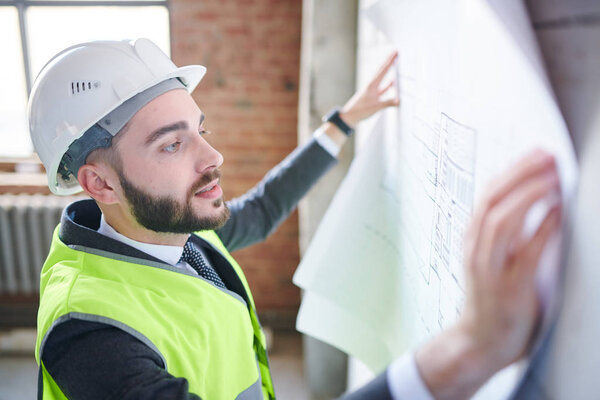 Young engineer in hardhat and uniform looking at sketch on blueprint while standing in front of wall of unfinished building