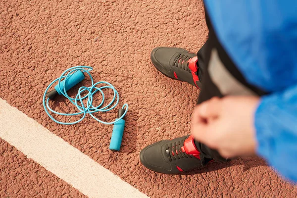 体育场赛道上的蓝色折叠跳绳概述 以及站在附近的年轻运动员的脚 — 图库照片