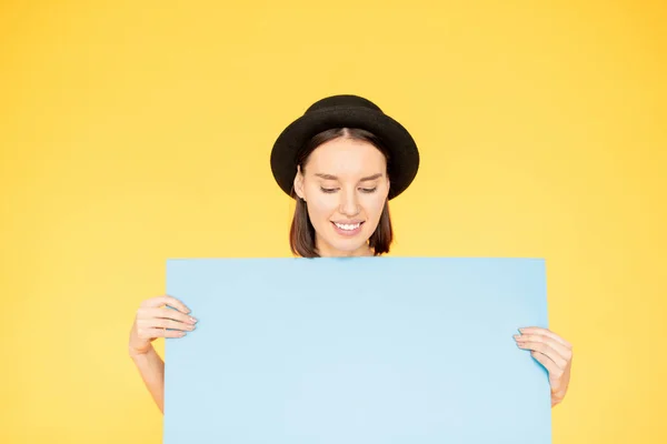 面带微笑 头戴黑色帽子的女性在黄色背景上做广告时 看着手中的蓝色床单 — 图库照片