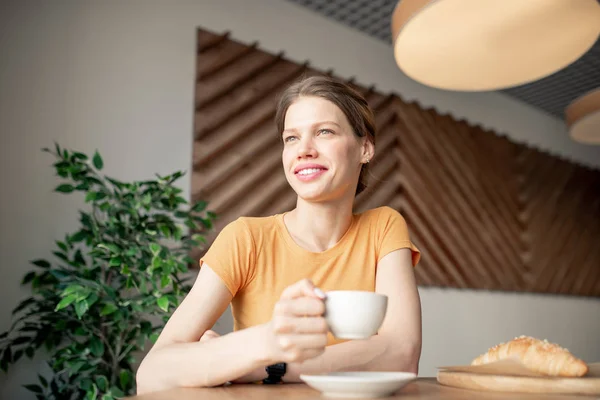 快乐的年轻休闲女人在T恤透过窗户看 而坐在小咖啡馆的桌子和有一杯美味的咖啡 — 图库照片