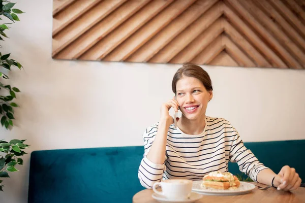 Young cheerful woman in casualwear talking by smartphone while sitting by table in cafe and having coffee and dessert