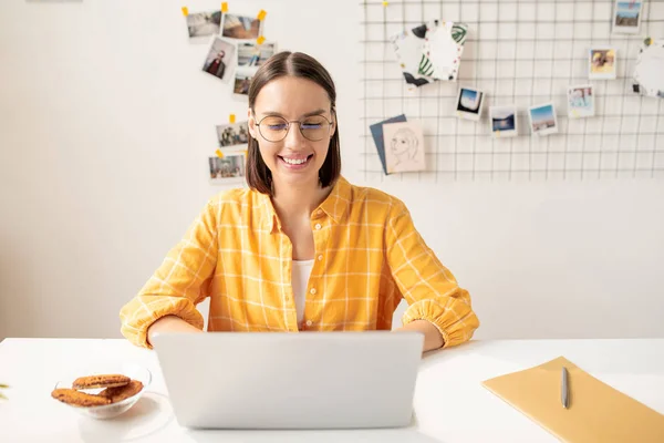 穿着便服的年轻而快乐的黑发女人坐在笔记本电脑前 看视频 在办公桌旁吃零食 — 图库照片