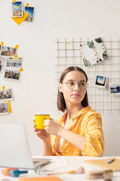 年轻而严肃的女商人或设计师 穿着黄色休闲衫 坐在笔记本电脑前的桌子旁边喝茶或喝咖啡 — 图库照片