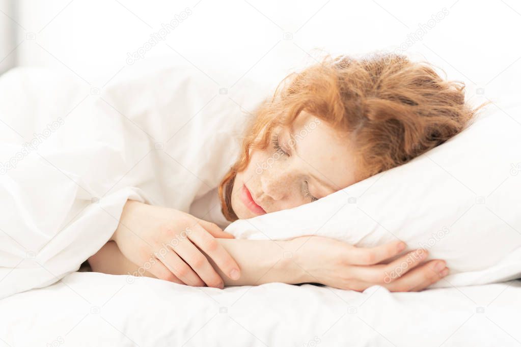Cute ginger-head girl with closed eyes lying on soft wite pillow under blanket or sheet before waking up in the morning