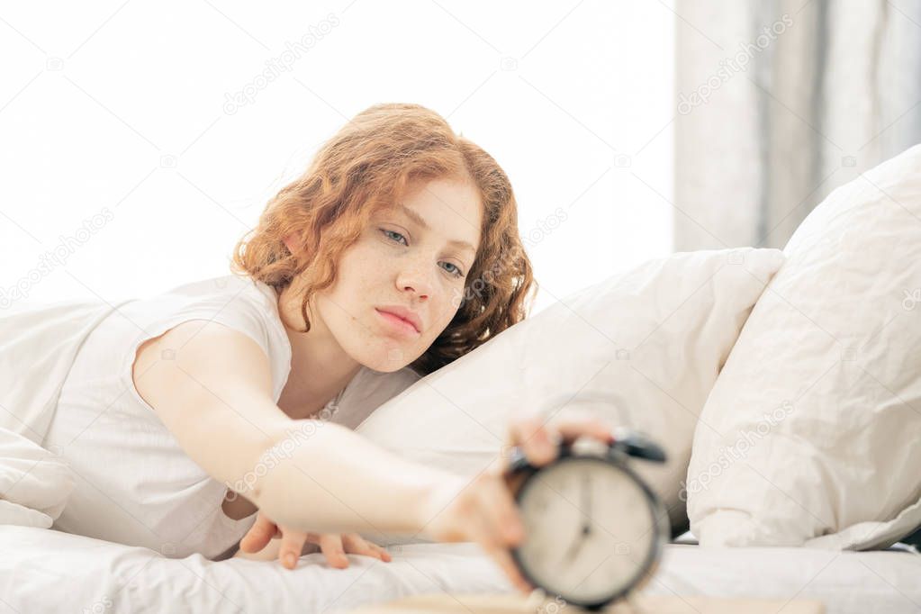 Displeased young woman lying on bed while stretching hand to turn off the alarm clock in the morning