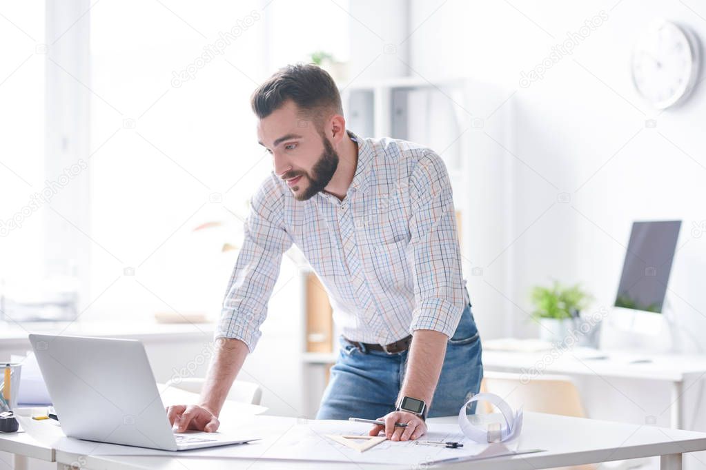Young confident businessman or architect in shirt and jeans looking at laptop display while bending over desk on working day