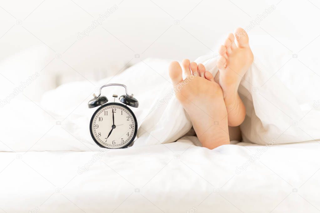 Alarm clock on bed next to bare female feet and soles stuck out of white blanket or sheet during sleep