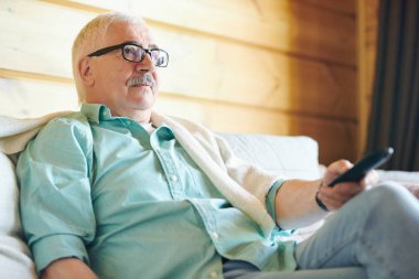 Onun kır evinde tv izlerken uzaktan kumanda kullanarak gözlük ve casualwear restful kıdemli adam