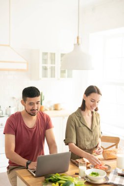Çağdaş genç adam mutfak masası tarafından dizüstü bilgisayar kullanarak eşi yanında duran ve kahvaltı için sebze sandviç yaparken