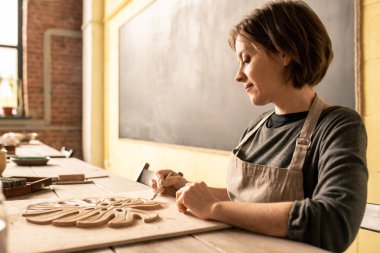 Yeni öğe üzerinde çalışırken yumuşak şeyler şekilleri kesmek için özel el aleti kullanarak genç kadın kil tasarımcısı
