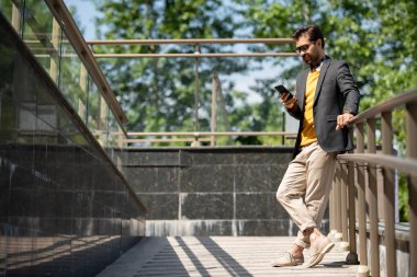 Bej kot pantolon genç rahat adam, sarı pullover ve açık korkuluklar tarafından akıllı telefon kaydırma koyu mavi ceket