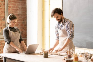 Apron genç adam yeni kil öğe üzerinde çalışırken kulaklık onun kadın meslektaşı toprak tasarımı hakkında online video izlerken