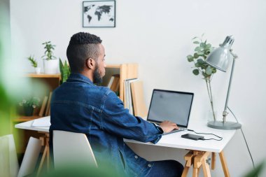 Kot ceketli genç rahat adam masa başında sandalyede oturan ve iş sırasında önünde dizüstü ekran bakarak