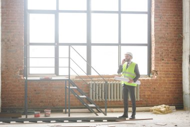 Bitmemiş binanın içinde dururken cep telefonu ile konuşan hardhat ve iş kıyafeti genç foreman veya müteahhit