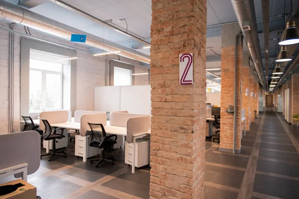 Interior of modern loft office open space with desks and swivel chairs, uncovered pipes on ceiling and long corridor with hanging simple lamps