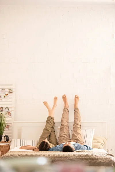 年轻的无忧无虑的赤脚约会在休闲服躺在床上 他们的凸起的腿对面的墙壁 而在家里放松 — 图库照片