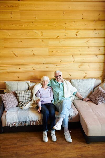 Affectionate senior couple in casualwear relaxing on sofa in their country house on background of wooden wall