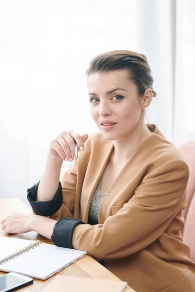 Portrait of content confident brunette business lady in beige jacket sitting at table and keeping list of tasks