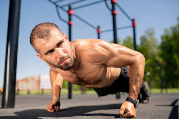 Young muscular bodybuilder standing on fists over ground while doing push-ups during workout outdoors