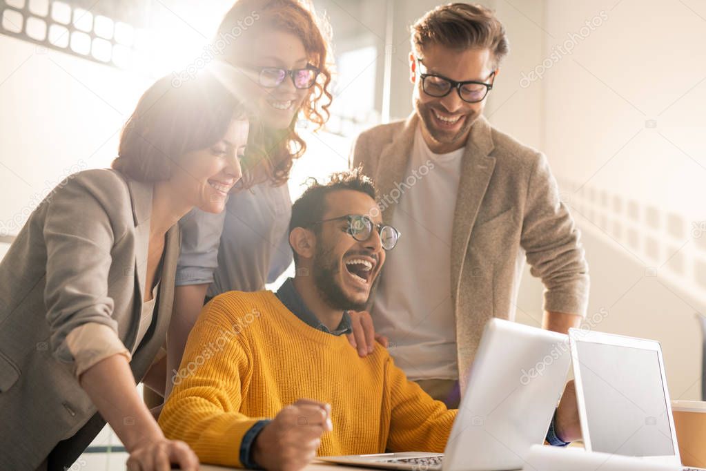 Group of cheerful excited young interracial colleagues laughing while reading internet news on laptop