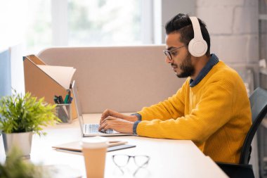 Dizüstü bilgisayar ile çalışırken sakallı düşünceli modern genç Arap pazarlamacı masada oturan ve kablosuz kulaklık müzik dinlerken