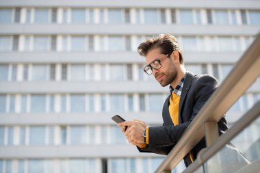 Gözlüklü mobil işadamı ve akıllı telefonunda akıllı telefondaki rahat okuma bildirimi, kentsel çevrede tırabzanlara yaslandı