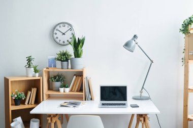 Masa, lamba, fotokopi defterleri, kitap ve bitkiler, gadget'lar ve duvarda saat ile raflar oluşan çağdaş ofis çalışanının işyeri