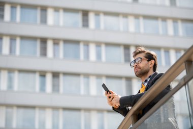 Kentsel çevrede ofis merkezinin arka planda banisters tarafından ayakta akıllı telefon ile pensive veya ilham genç bin yıl