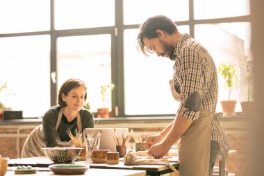 Apronda genç erkek çömlekçi masanın yanında duruyor ve iş arkadaşı dizüstü bilgisayarın önünde ağ yaparken kil eşyalardan biri üzerinde çalışıyor