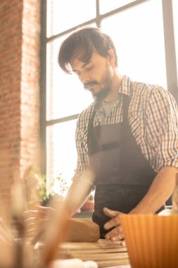 Atölyede çalışma sürecinde işyerinin yanında dururken önlükteki genç çömlekçi kil parçasına bakıyor