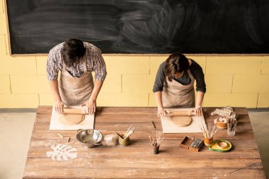 Önlükler içinde masanın yanında duran ve oklavalarla kil yuvarlanan iki genç çağdaş serbest meslek sahibi çömlekçi görünümü