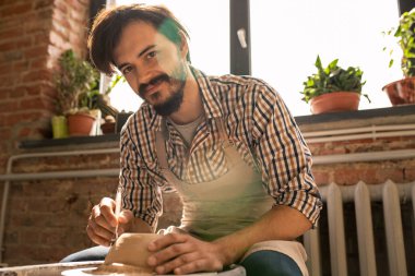 Kendi atölyesinde yeni kil öğe üzerinde çalışırken size bakarak iş elbisesi genç başarılı çömlekçi