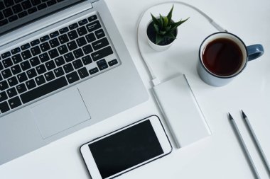 Dizüstü tuş takımı, akıllı telefon, kahve veya çay ile kupa, küçük saksı yeşil bitki, iki kalem ve masa üzerinde pil şarj görünümü