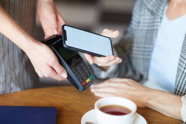 Genç garson masa üzerinde elektronik ödeme makinesi tutarken müşterilerinden biri kahve için ödeme yapmak için akıllı telefonda mobil uygulamayı kullanırken