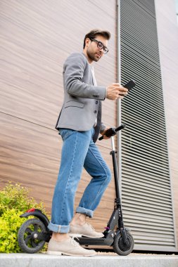 Mobil genç adam akıllı rahat onun scooter modern binanın duvar tarafından ayakta ve akıllı telefon kaydırma