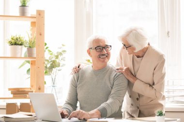 Yanında duran karısıyla konuşurken dizüstü bilgisayarın önünde masa başında oturan rahat giyimli mutlu yaşlı adam