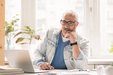 Ofiste dizüstü bilgisayar önünde masa başında otururken size bakarak casualwear olgun dalgın işadamı