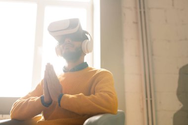 Sanal gerçeklik kulaklık meditasyon yaparken sakallı sakin genç Arap adam koltukta oturan ve avuç içi birleştirerek