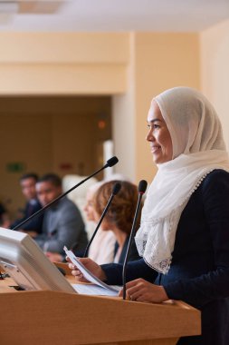 Oldukça genç gülümseyen Müslüman kadın konuşmacı tribün tarafından ayakta ve konferansta yabancı meslektaşları için rapor yapma