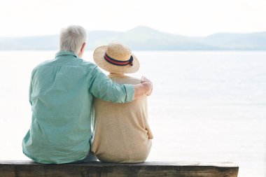 Yaz günü su kenarında dinlenirken göle bakan rahat giyimli sakin yaşlı çiftin arka görünümü