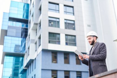 Modern bina açık havada ayakta iken touchpad kullanarak formalwear ve koruyucu kask genç sakallı mimar