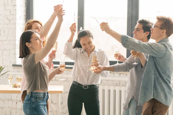 ホームパーティーで踊りながらベンガルライトで手を上げるシャンパンを持つ興奮した若い友人のグループ — ストック写真