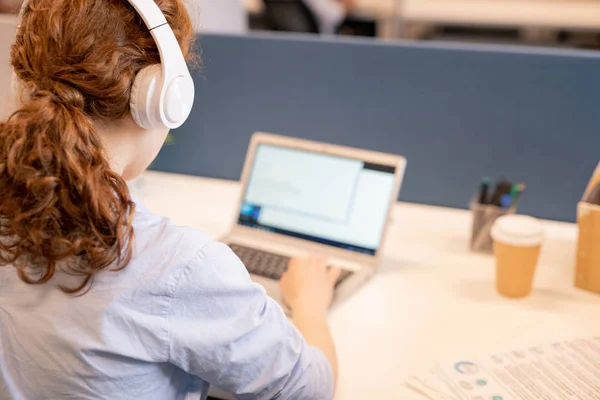 Rear view of young redhead businesswoman with ponytail listening to music in headphones while working with text file on laptop