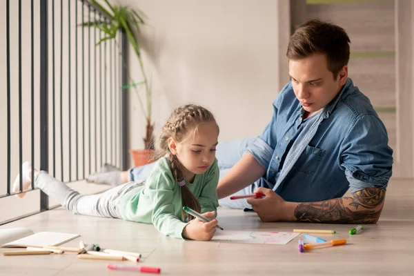 床に横たわり 娘にアドバイスを与えるデニムシャツの深刻なヒップスター若い父親 自宅で父親と一緒に絵を描く小さな女の子 — ストック写真