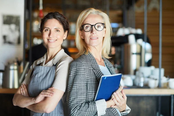 Two successful female entrepreneurs standing in front of camera on background of bar counter in modern cafe or restaurant