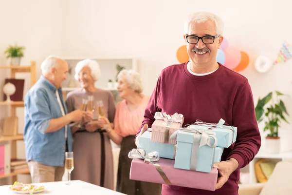 Joyful mature man with several giftboxes standing in front of camera while enjoying party with friends